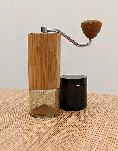 Handkaffeemühle mit Holzverkleidung, Kurbel und gläsernem Auffangbehälter neben einem dunklen Vorratsglas.