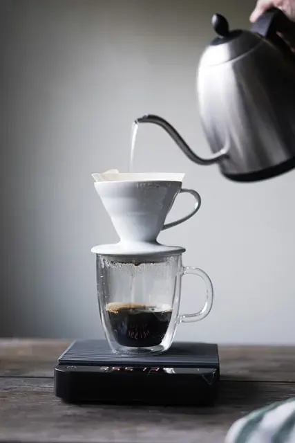 Aufguss mit einem Schwanenhalskessel in einen weißen V60-Handfilter über einer Glastasse auf einer Digitalwaage.