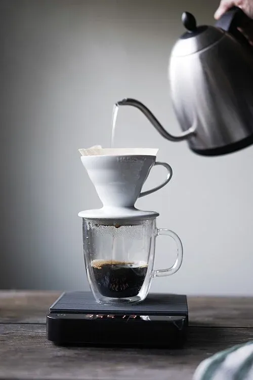 Aufguss mit einem Schwanenhalskessel in einen weißen V60-Handfilter über einer Glastasse auf einer Digitalwaage.