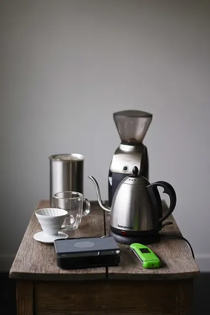 Kaffeezubehör mit digitaler Waage im Vordergrund, Handfilter, Glastasse, Schwanenhalskessel und elektrischer Mühle.