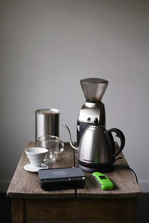 Kaffeezubehör mit digitaler Waage im Vordergrund, Handfilter, Glastasse, Schwanenhalskessel und elektrischer Mühle.