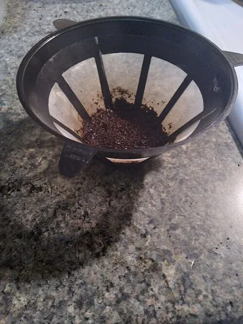 Herausgenommener wiederverwendbarer Kaffeefilter mit schwarzem Rahmen, feinem Gewebe und Kaffeerückständen.