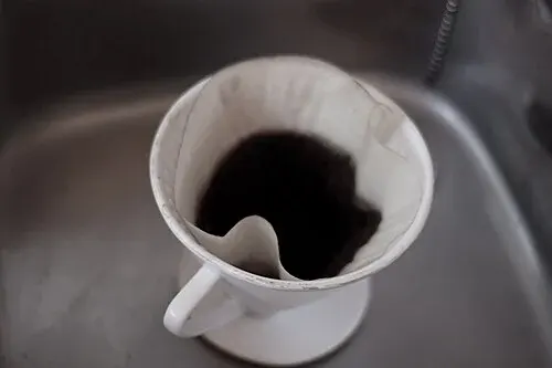 Weißer Handfilter mit Filterpapier und benutztem Kaffeemehl auf einer Küchenspüle.