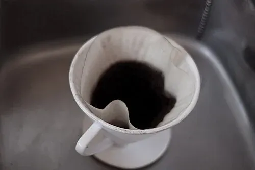 Weißer Handfilter mit Filterpapier und benutztem Kaffeemehl auf einer Küchenspüle.