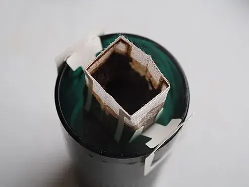 Geöffneter Kaffee-Portionsfilter mit nassem Kaffeemehl, der mit Papphaltern auf einer Tasse sitzt.