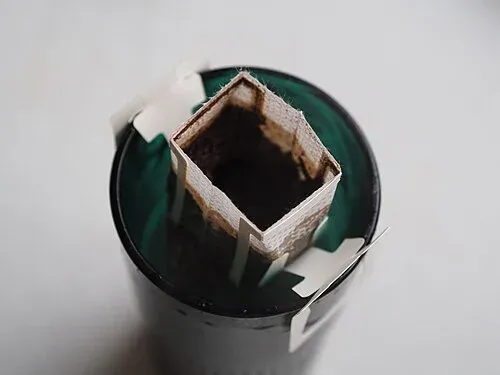 Geöffneter Kaffee-Portionsfilter mit nassem Kaffeemehl, der mit Papphaltern auf einer Tasse sitzt.