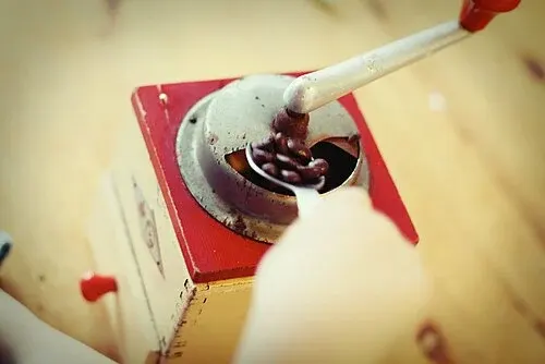 Handkaffeemühle mit rot-weiß lackiertem Gehäuse, Kurbel und Bohnen im Einfüllbereich.