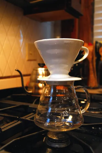 Hario-V60-Handfilter auf einem Glasserver; im Hintergrund steht ein Buono-Kessel.