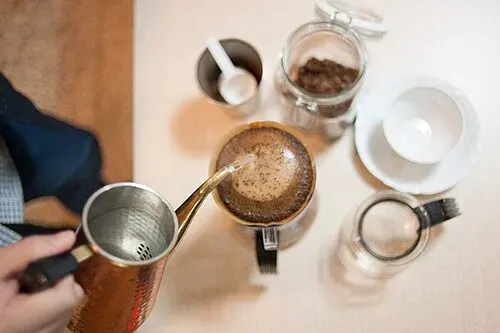 Kaffeeaufguss von oben mit schmaler Kesseltülle, gefülltem Handfilter, Bohnenvorrat, Messlöffel und Tasse.