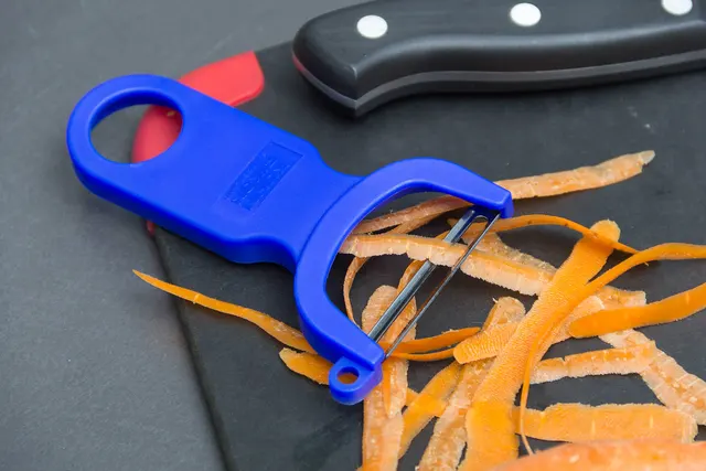 Blauer Y-Schäler mit Karottenschalen auf einem Schneidbrett neben einem Messergriff