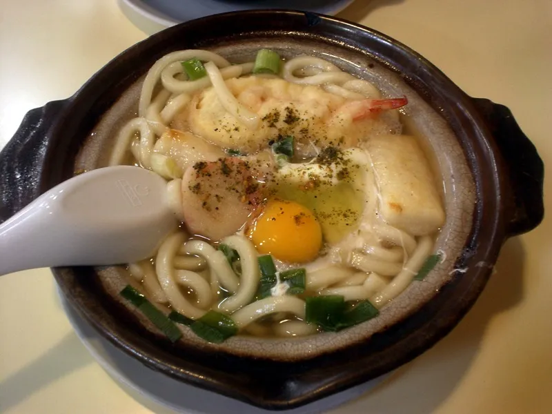 Udon-Nudeln mit Ei und Garnele in einem dunklen Keramiktopf mit zwei Griffen