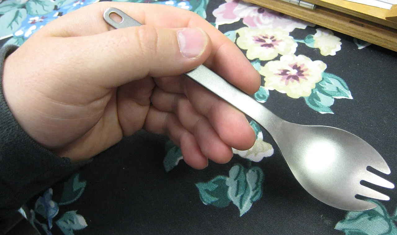 Una mano regge uno spork in titanio, utensile che combina cucchiaio e forchetta.