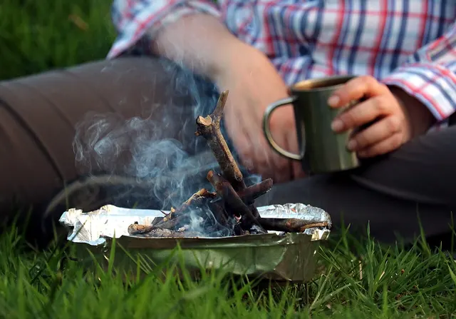 Una persona con una tazza in acciaio accanto a un piccolo fuoco da campo