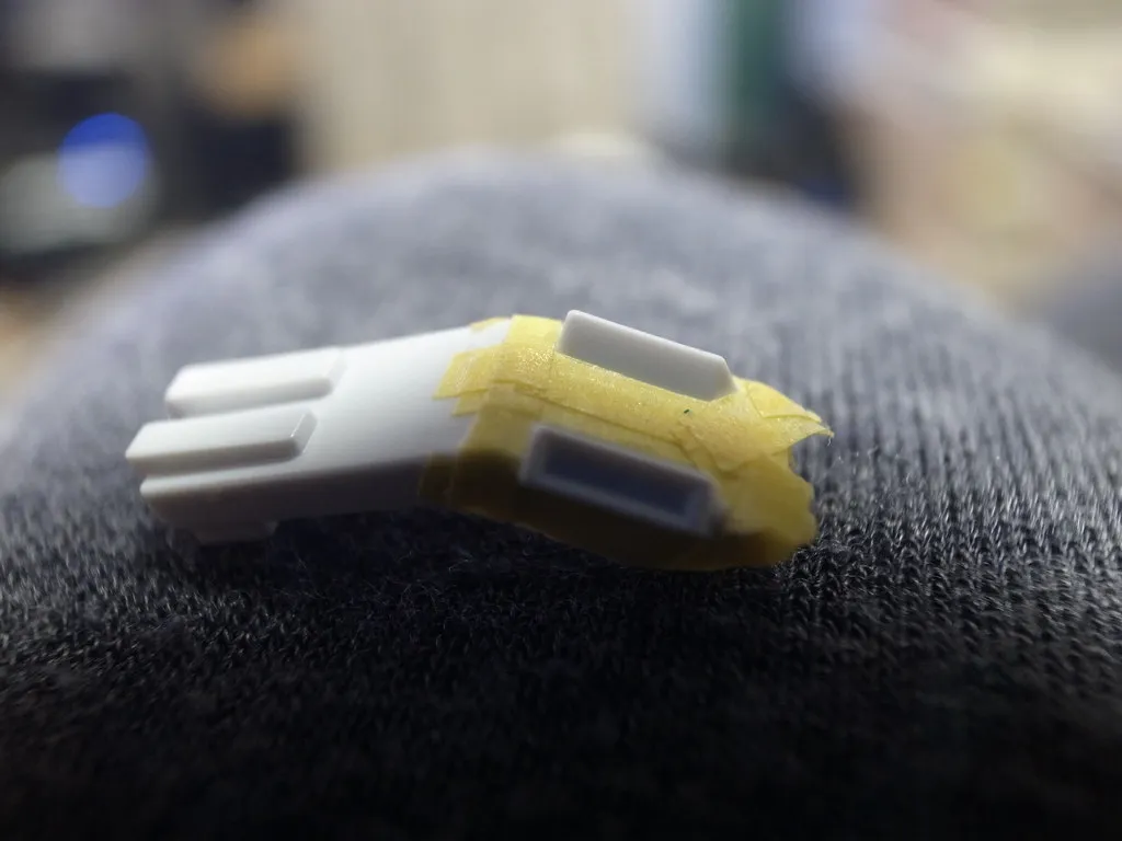 En liten plastdel som delvis täckts med gul maskeringstejp.