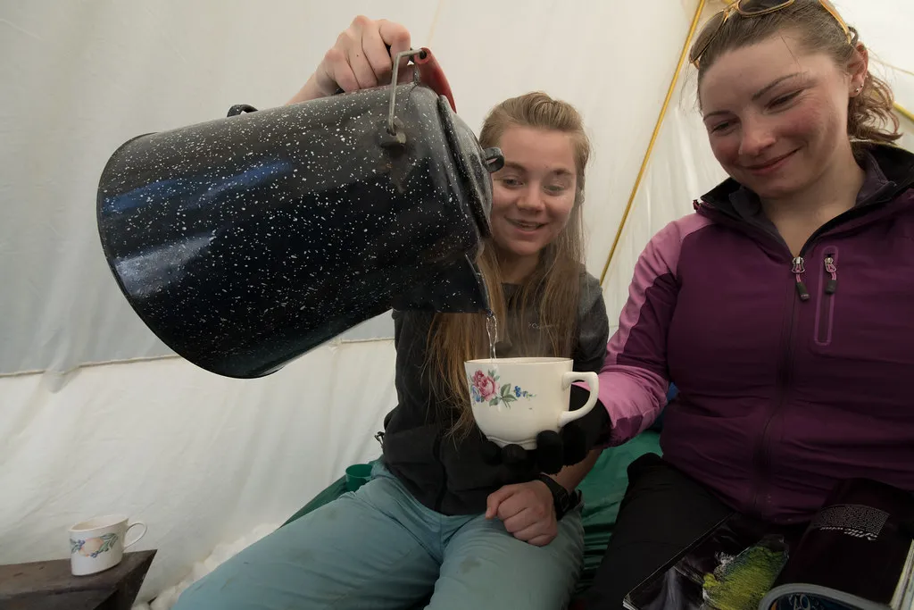 Zwei Camperinnen gießen im Zelt Tee aus einer großen Kanne in einen Becher