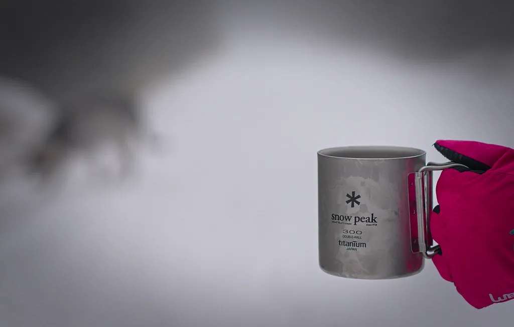 Snow-Peak-Titanbecher mit Henkel neben einem farbigen Textilteil
