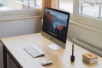 Monitorfuß, Tastatur und Maus auf einem schmalen Schreibtisch.